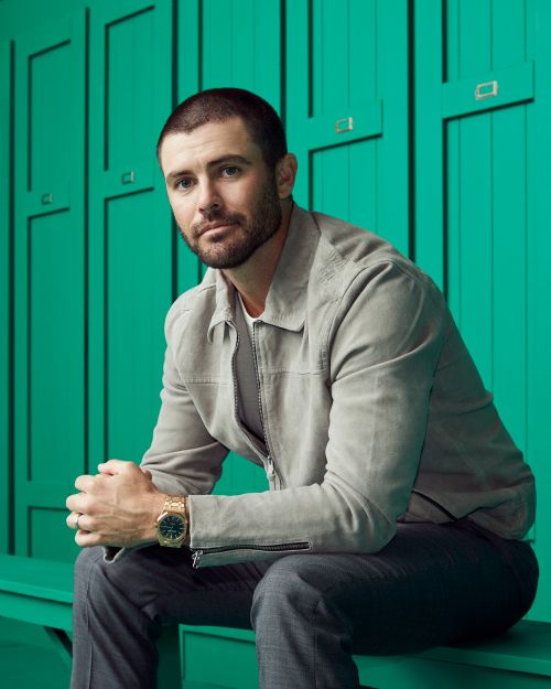 An adult man with a beard and buzz cut is sitting on a bench wearing a light gray jacket, gray pants, and gold watch, posing indoors against a green wall.