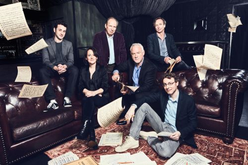 A group of six people seated on a leather couch surrounded by scattered sheet music in a dimly lit, indoor setting.