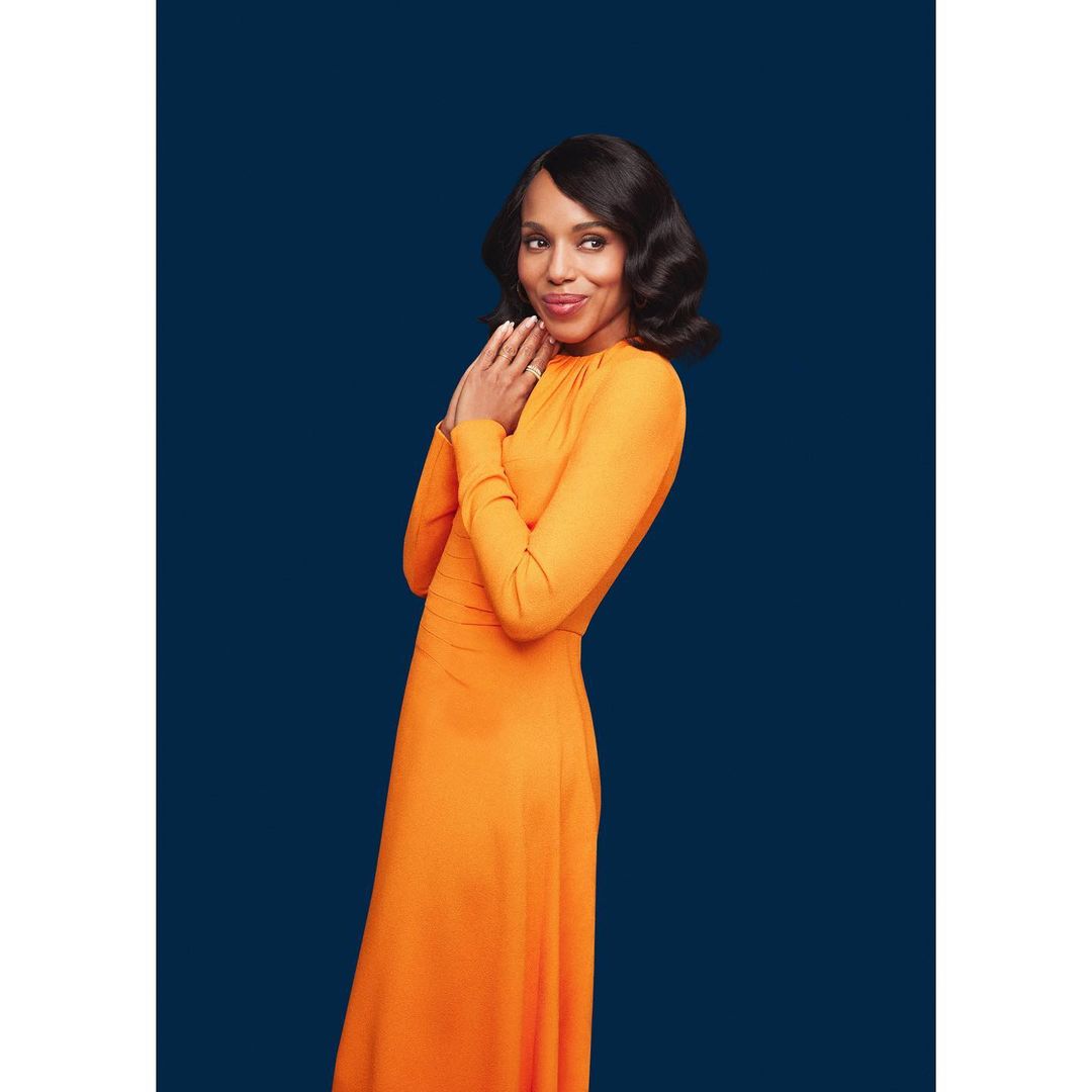 Portrait of an adult woman in an orange gown posing indoors against a navy background with hands near her face and dark wavy hair.