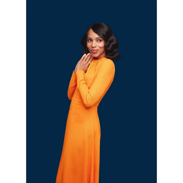 Portrait of an adult woman in an orange gown posing indoors against a navy background with hands near her face and dark wavy hair.