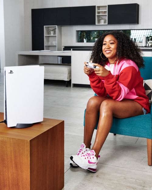 Young woman smiles while playing a video game on a PlayStation 5, sitting in a teal chair in a modern home interior.