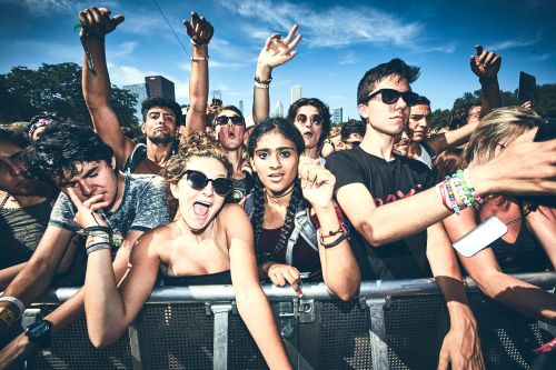 Enthusiastic crowd of diverse young adults at an outdoor music festival in the city, cheering with raised hands under a bright blue sky.