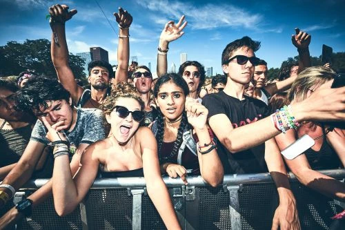 Enthusiastic crowd of diverse young adults at an outdoor music festival in the city, cheering with raised hands under a bright blue sky.
