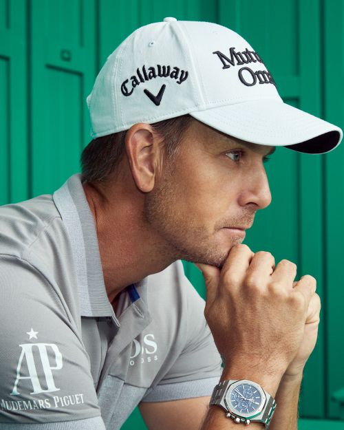 Close-up of a man in a Callaway golf cap and grey polo shirt with sponsor logos, hand on chin, against a green background.