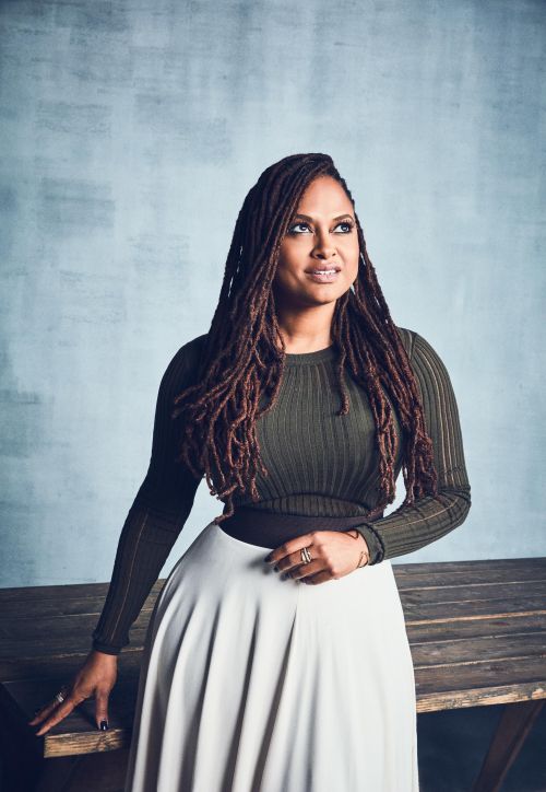 Portrait of an adult woman with dreadlocks wearing a green long-sleeve shirt and white skirt, posing against a light-blue wall indoors.