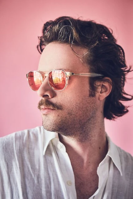 Portrait of a man with dark hair and a mustache wearing mirrored sunglasses and a white linen shirt against a light pink background.