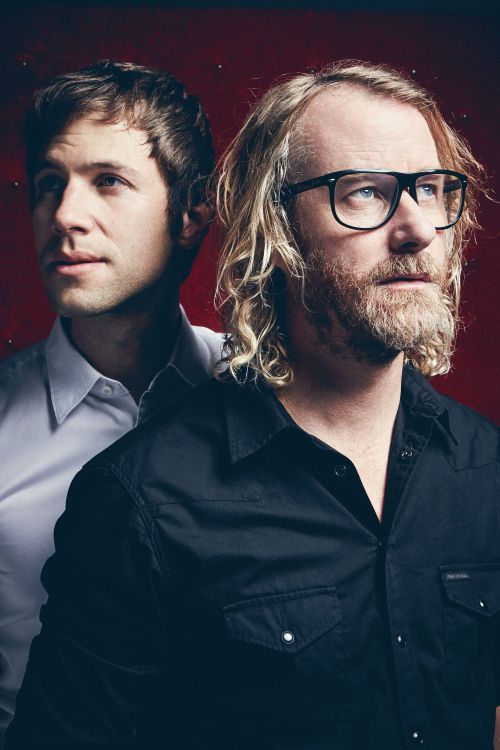 Portrait of two men, one wearing thick black glasses and a black shirt, photographed by Koury Angelo against a textured red background.