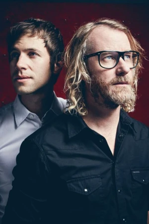 Portrait of two men, one wearing thick black glasses and a black shirt, photographed by Koury Angelo against a textured red background.