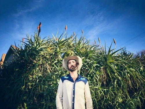 Man in cowboy hat stands in front of large aloe plants beneath a blue sky. He wears a Western-style shirt in outdoor setting.