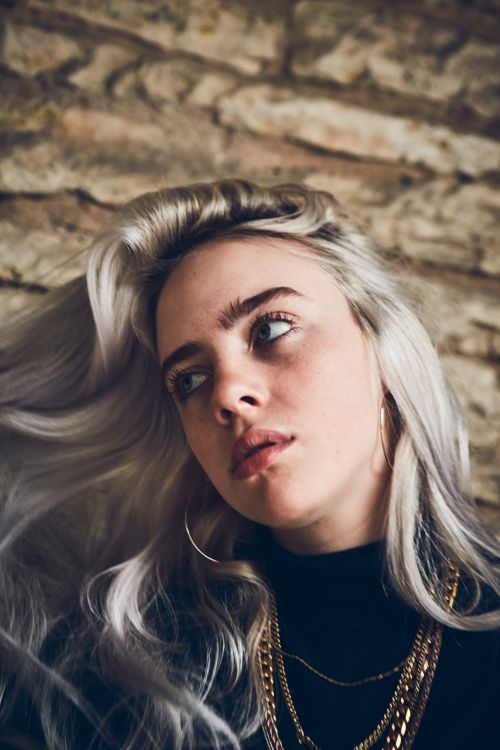 Portrait of a woman with platinum blonde hair, wearing a black turtleneck and gold chains, against a textured beige background.