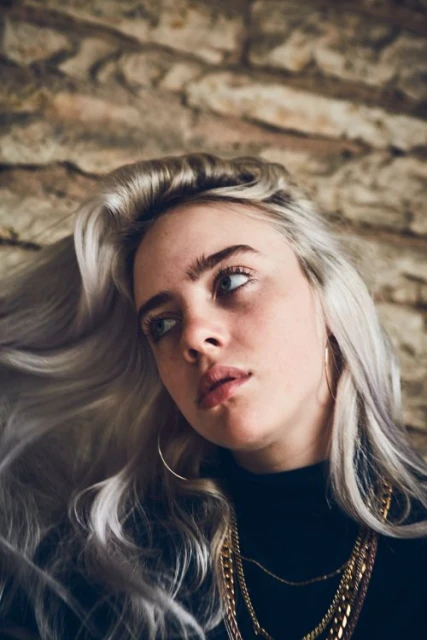 Portrait of a woman with platinum blonde hair, wearing a black turtleneck and gold chains, against a textured beige background.