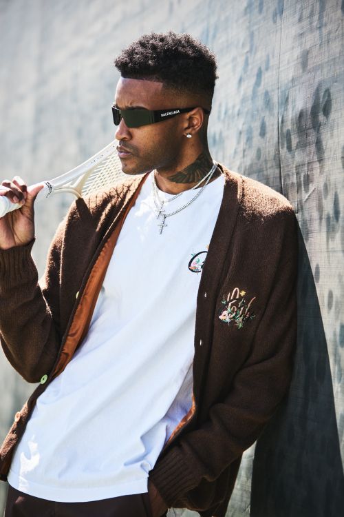 Stylish adult male poses in brown cardigan with sunglasses and jewelry, holding tennis racket against textured background in an outdoor setting.