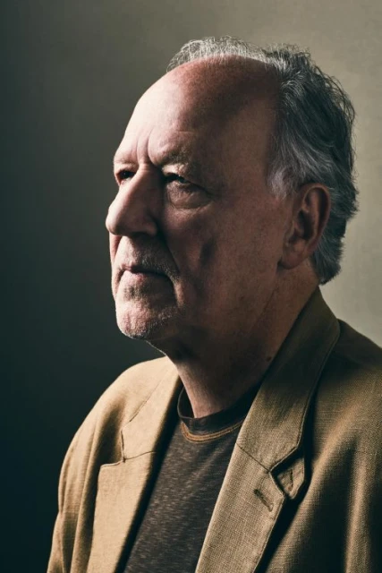 Portrait of an older man with grey hair wearing a tan blazer and brown shirt against a dark background in a softly lit indoor setting.