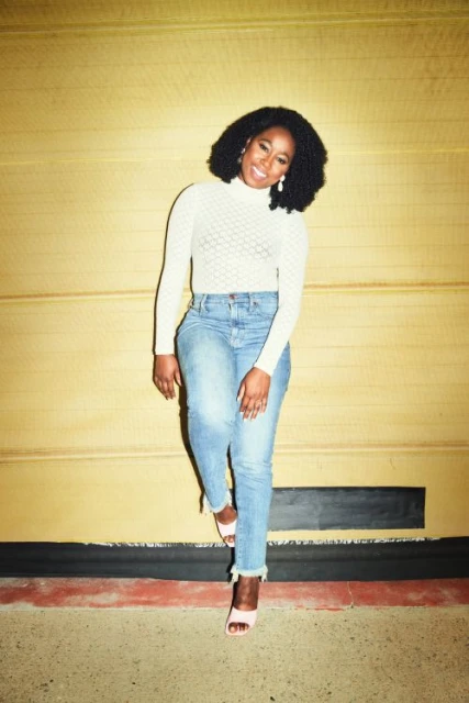 Woman in light jeans and a white shirt poses against a textured yellow wall indoors, casting a cheerful and casual vibe.