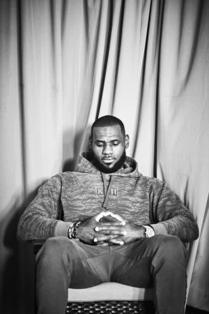 LeBron James seated in an armchair wearing a patterned hoodie with clasped hands against a heavy draped curtain background.