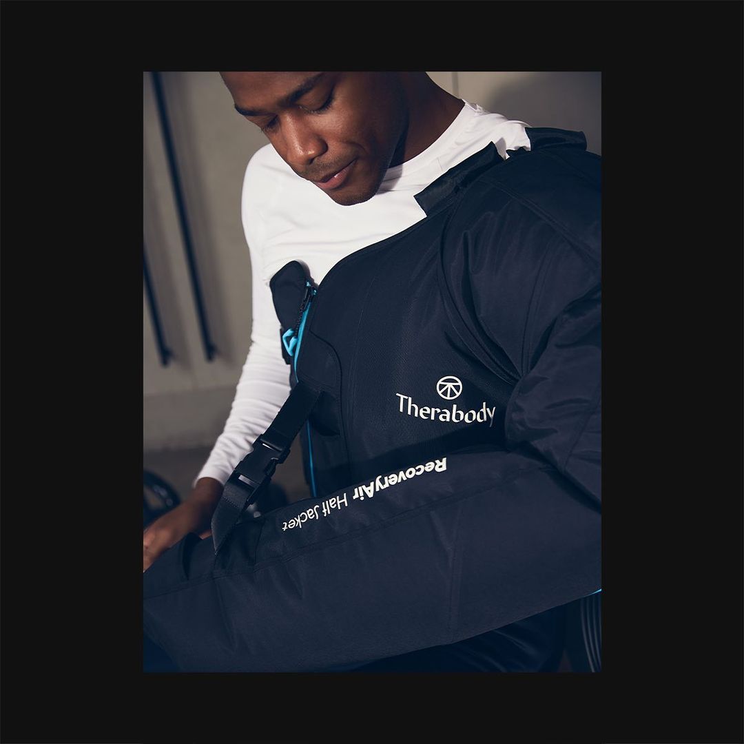 An adult male wearing a Therabody RecoveryAir Half Jacket, designed for post-workout recovery, in a neutral indoor environment.