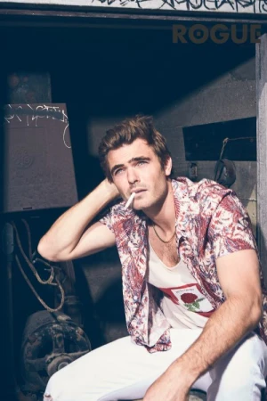 Man in patterned short-sleeve shirt with cigarette in mouth sitting outdoors by a dark wall