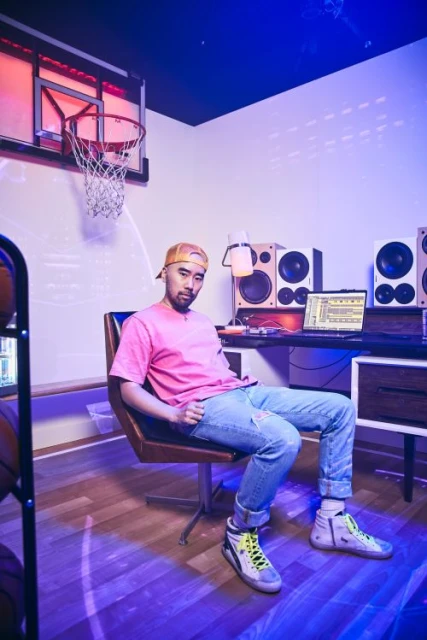 Man sits in a chair in a room with a basketball hoop, computer, and speakers. He wears a pink t-shirt, jeans, and sneakers.