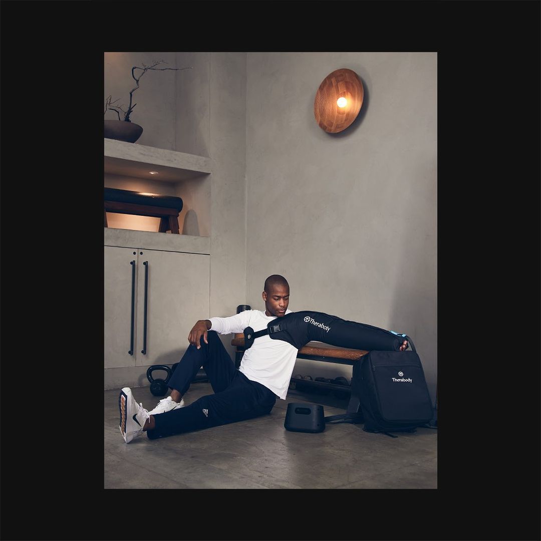 Black man using a Therabody recovery system on arm. He is wearing athletic clothing in a modern, minimal indoor setting with weights.