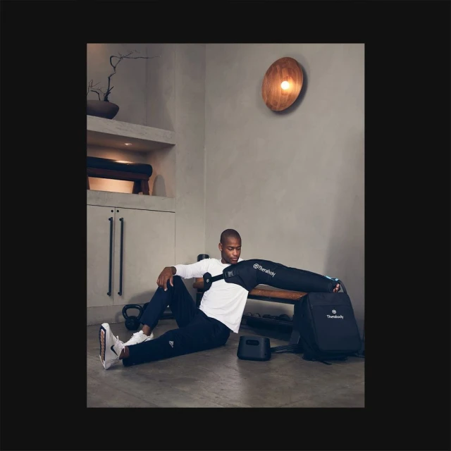 Black man using a Therabody recovery system on arm. He is wearing athletic clothing in a modern, minimal indoor setting with weights.