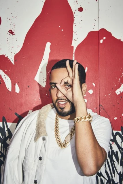 French Montana poses in front of an abstract painted wall, wearing a white jacket, t-shirt, and jewelry, making an "OK" sign with his fingers.