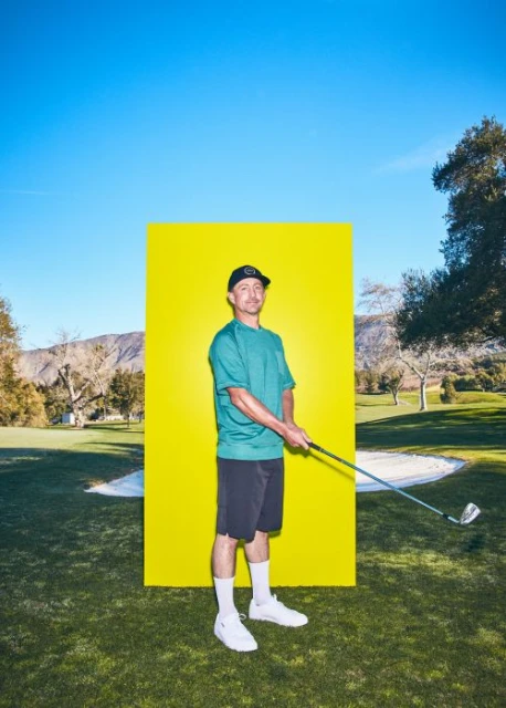 An adult male golfer wearing a teal shirt and black shorts poses with a golf club against a yellow backdrop in a sunny outdoor setting.
