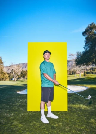 An adult male golfer wearing a teal shirt and black shorts poses with a golf club against a yellow backdrop in a sunny outdoor setting.