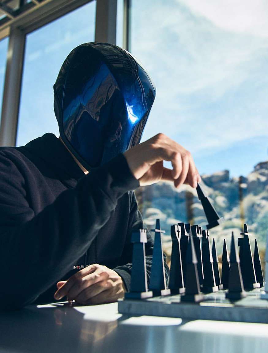 Person wearing a blue geometric mask playing chess. Chess pieces are lined up on a marble table near a bright window.