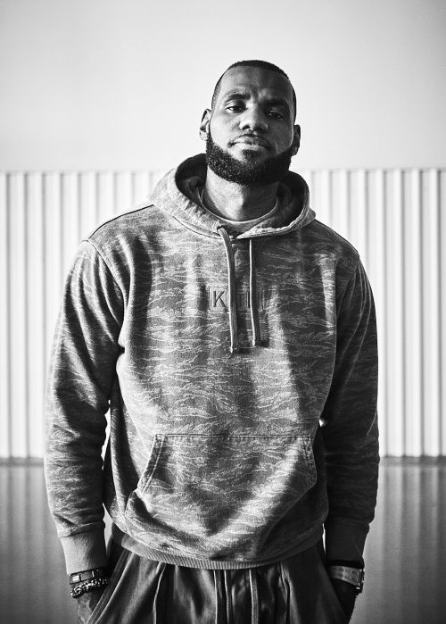 Portrait of LeBron James in a gray camo hoodie, standing indoors with a neutral expression and hands down by his side in soft lighting.