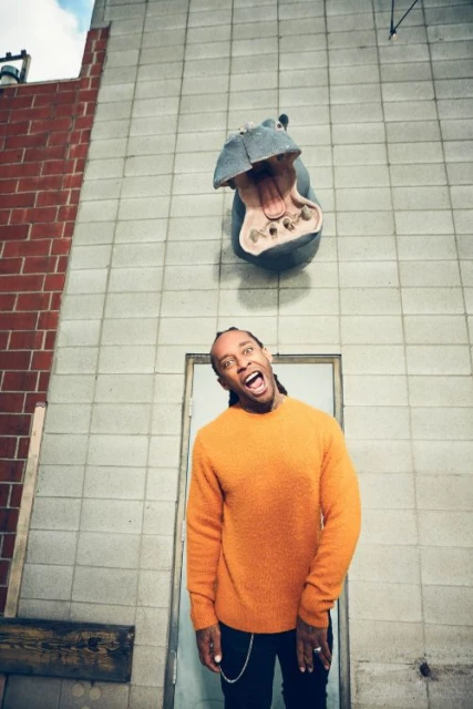 Adult man with an open mouth poses in front of a hippo head sculpture on a building. He wears an orange sweater and black pants.