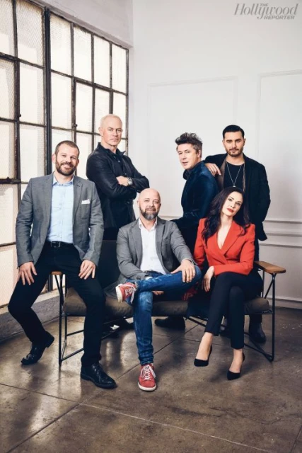 Group portrait of six people, including five men and one woman, seated and standing in a photo studio with natural window light.