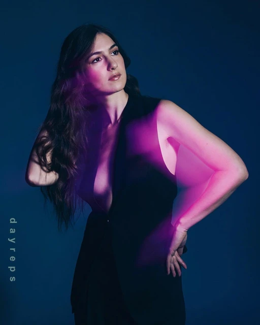 Portrait of a brunette woman with long hair in a sleeveless black dress, posing against a blue background with pink lighting effects.