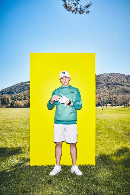 A man in golf attire poses in front of a yellow panel on a grassy lawn with mountains in the background under a clear blue sky.