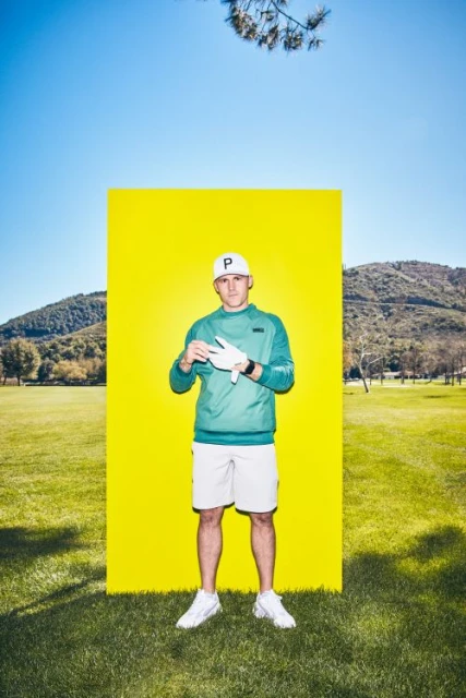 A man in golf attire poses in front of a yellow panel on a grassy lawn with mountains in the background under a clear blue sky.