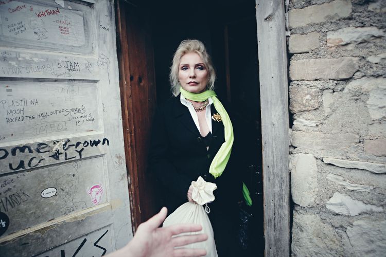 Blondie singer Debbie Harry in a doorway, wearing a black blazer, holding a bag, with graffiti-covered door and a stone wall.