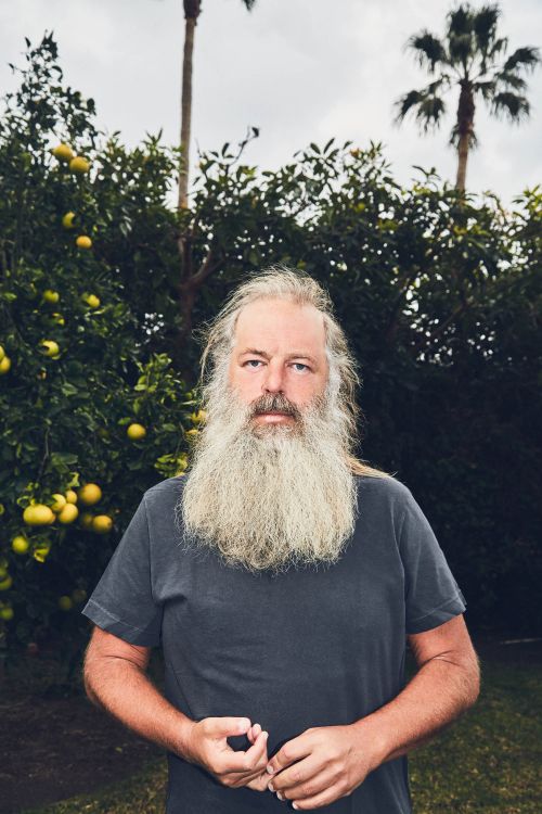 Portrait of a mature man with a long beard in a gray t-shirt, standing outdoors in front of citrus trees and palm trees on a cloudy day.