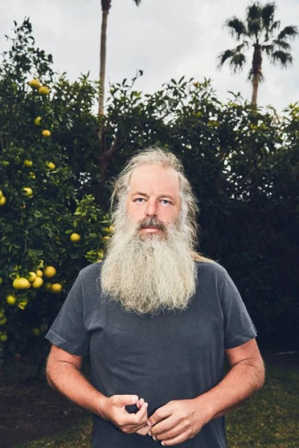 Portrait of a mature man with a long beard in a gray t-shirt, standing outdoors in front of citrus trees and palm trees on a cloudy day.