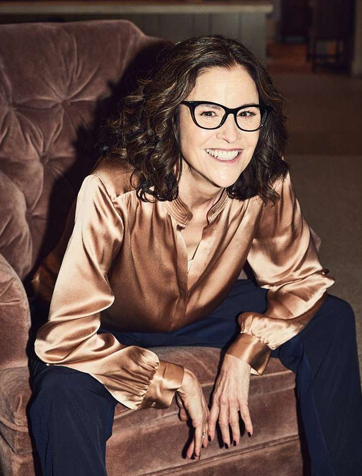 Portrait of a smiling middle-aged woman wearing glasses, a satin blouse, and dark pants, sitting in a plush armchair indoors.