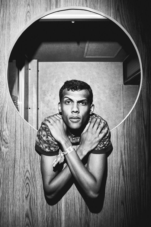 Monochrome portrait of a man looking through a circular cutout in a wood-paneled wall with arms crossed over his shoulders.
