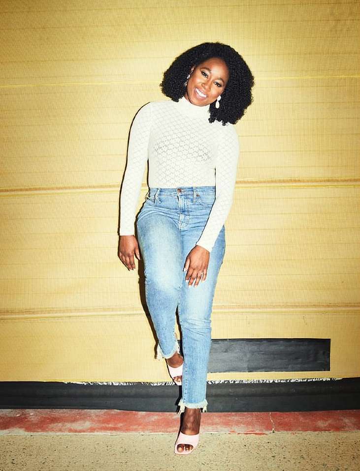 Smiling Black woman in long-sleeve top and jeans poses against a gold wall, showcasing a stylish indoor fashion portrait.