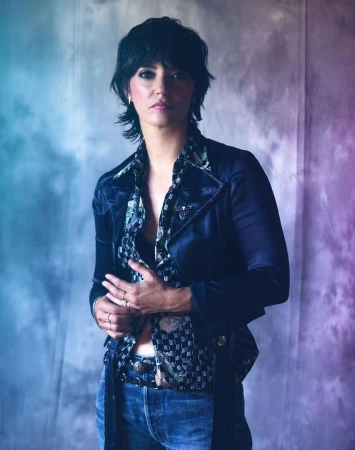 Portrait of a woman with short dark hair wearing a patterned shirt and blue jeans against a mottled blue backdrop