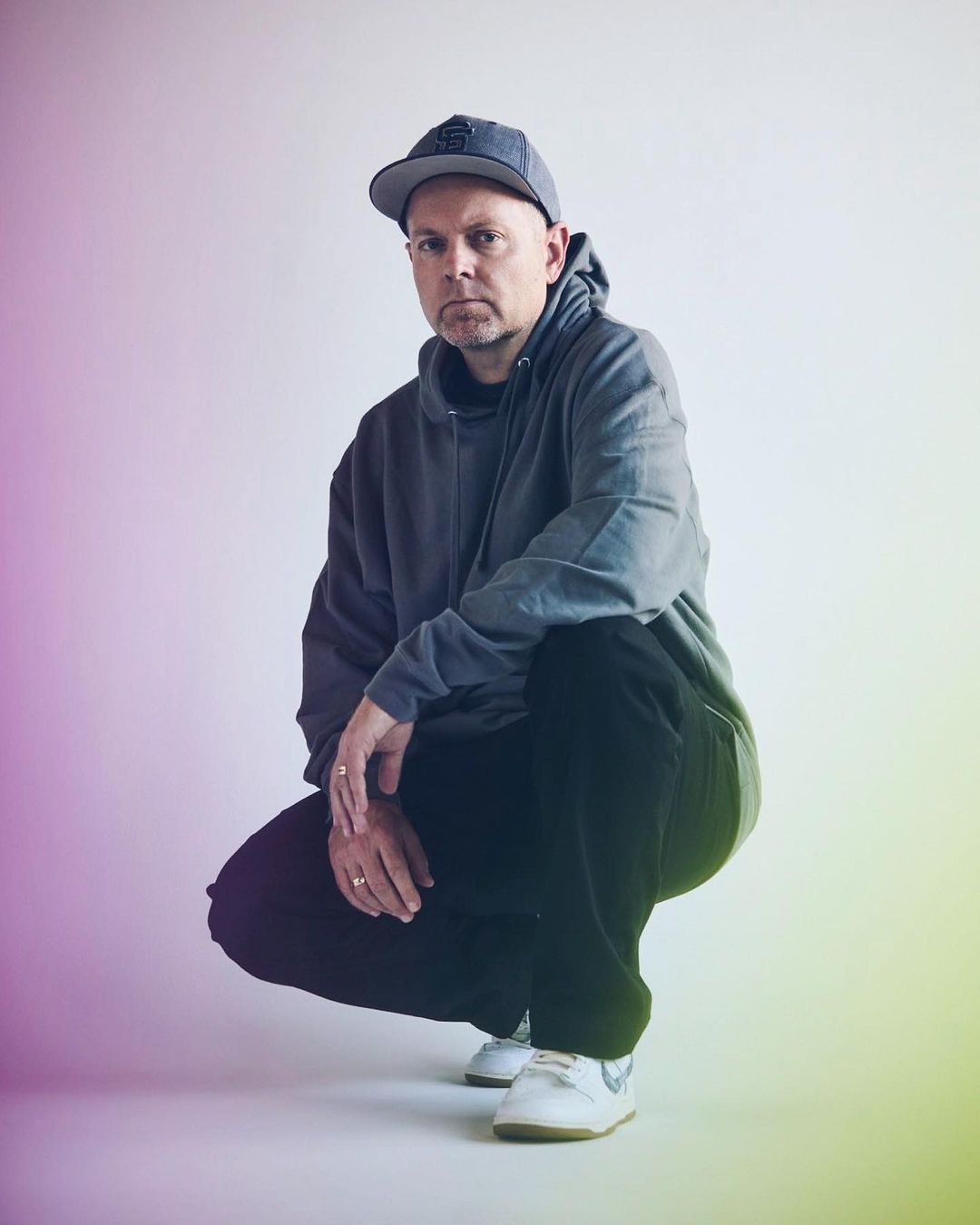 Man crouching wearing a grey hoodie, baseball cap, black trousers, and white sneakers against a gradient background.