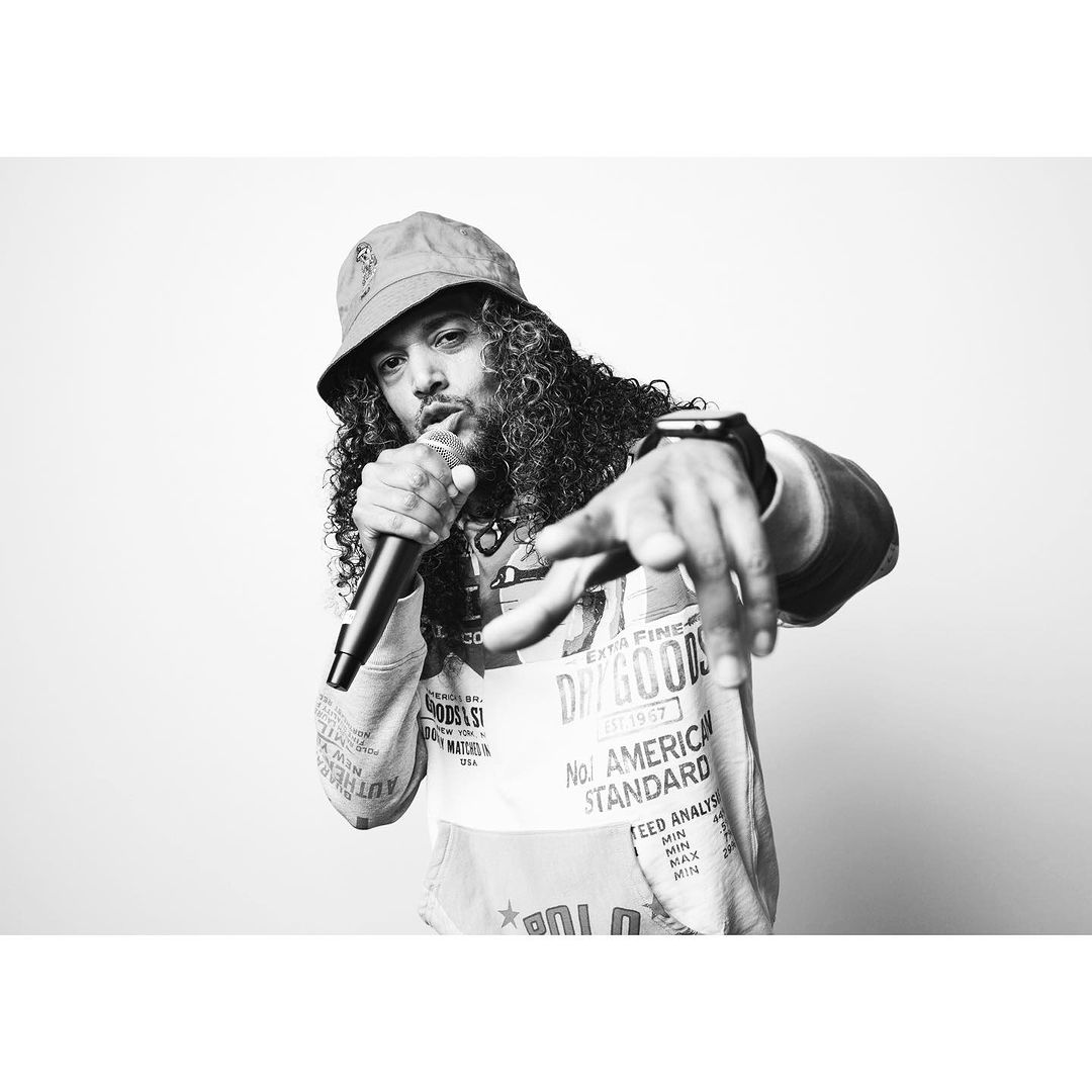 Black and white studio shot of a rapper holding a microphone, wearing a bucket hat and patterned sweatshirt with his hand reaching out.
