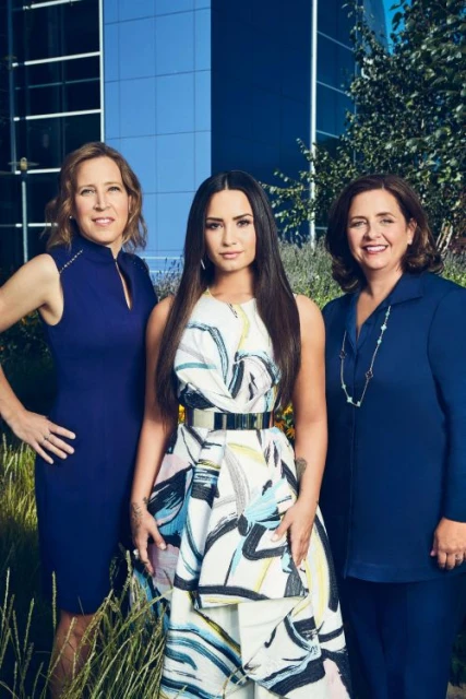 Three women stand outdoors in front of a modern building, bathed in natural light. The central woman wears a striking patterned dress.