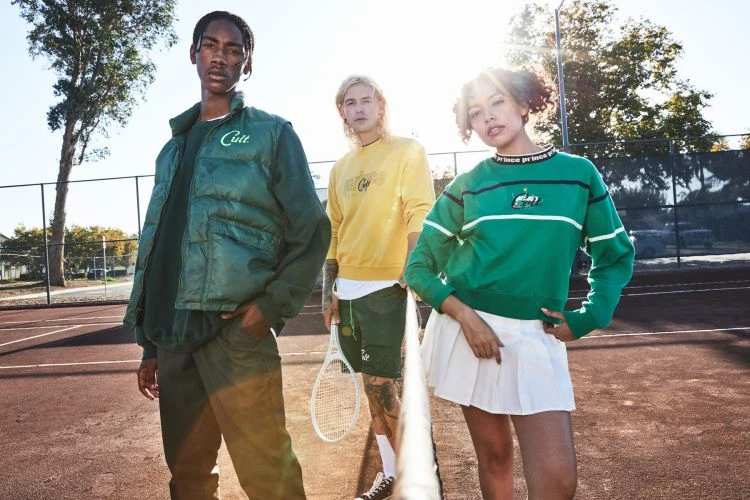 Three fashionable young adults stand on a tennis court, wearing green and yellow Prince x Cult collab streetwear, bathed in bright sunlight.