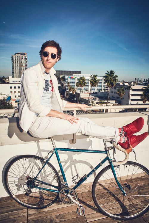 A man sits on a blue bicycle against an urban backdrop of Los Angeles buildings and palm trees under a bright blue daytime sky.