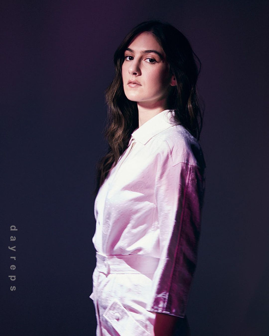 Portrait of a young adult woman with long brown hair wearing a pink button-down top and matching pants in a studio with dark lighting.