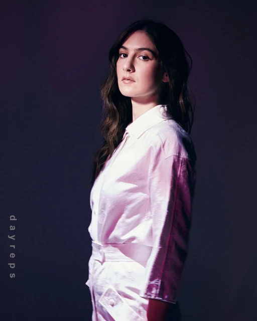Portrait of a young adult woman with long brown hair wearing a pink button-down top and matching pants in a studio with dark lighting.