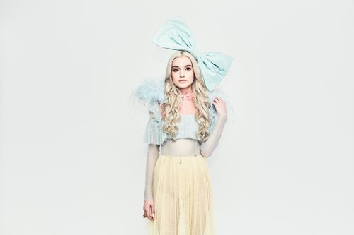 Portrait of a woman with blonde wavy hair, wearing a blue top, yellow skirt, and a large blue bow on her head in a studio setting.