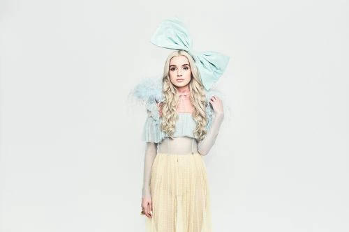 Portrait of a woman with blonde wavy hair, wearing a blue top, yellow skirt, and a large blue bow on her head in a studio setting.
